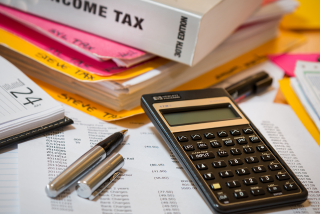 Mesa de trabalho com calculadora, canetas, documentos fiscais, calendário e um livro sobre imposto de renda.