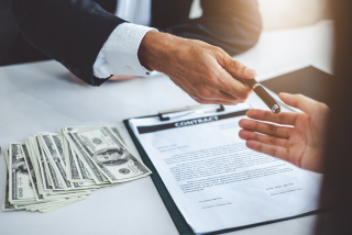 Pessoa entregando uma caneta para assinatura de contrato sobre a mesa, ao lado de notas de dólar