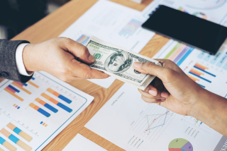Duas pessoas trocam um maço de notas de dólar americano sobre uma mesa de escritório coberta por gráficos e relatórios financeiros coloridos.