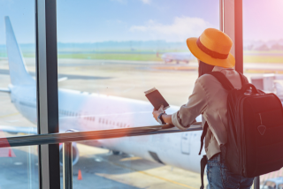 Uma pessoa vista de costas observa um avião estacionado na pista através das grandes janelas de um aeroporto. Ela usa um chapéu amarelo, mochila nas costas e segura o passaporte junto à passagem. A luz suave do sol entra pelas janelas.