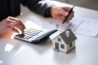 Pessoa utilizando uma calculadora e segurando uma caneta enquanto analisa documentos sobre uma mesa. Em primeiro plano, há uma pequena maquete de casa.