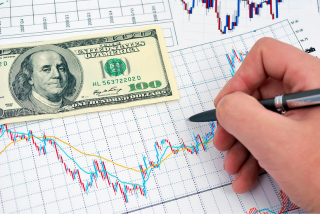 Gráfico financeiro com linhas de variação cambial. Uma mão segura uma caneta, com uma nota de 100 dólares posicionada sobre o papel.