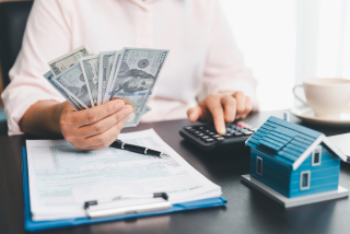 Pessoa sentada à mesa segurando várias cédulas de dólares enquanto usa uma calculadora com a outra mão. Sobre a mesa há documentos, uma caneta e uma pequena maquete de uma casa azul.